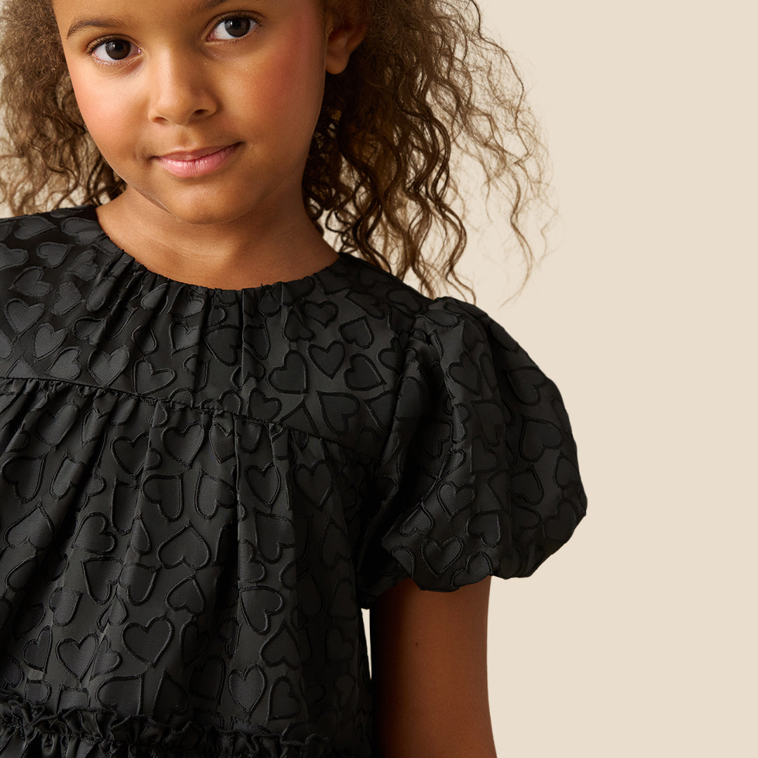 A young girl with curly brown hair wearing a black short-sleeve dress with a textured pattern, photographed against a solid beige background.