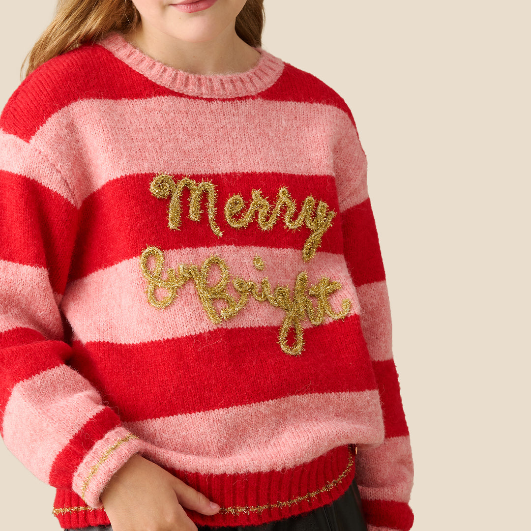 A young girl wearing a fuzzy knit sweater with red and pink horizontal stripes, featuring 'Merry Night' in gold glitter script across the chest, paired with a black faux-leather mini skirt, set against a solid beige background.