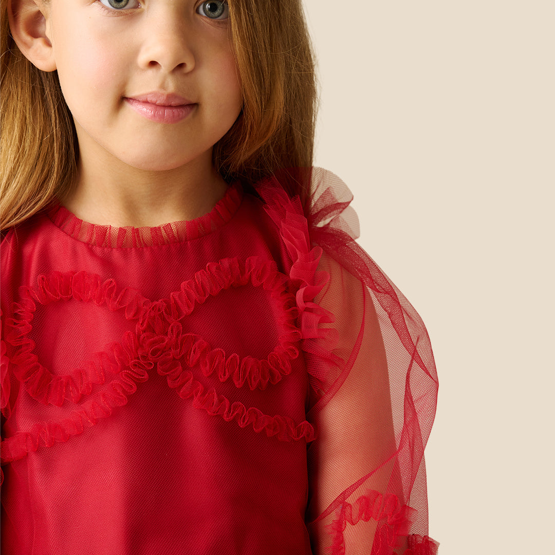 A young girl with blonde hair wearing a red tulle dress with red lace appliques and sheer flutter sleeves, standing against a solid beige background.