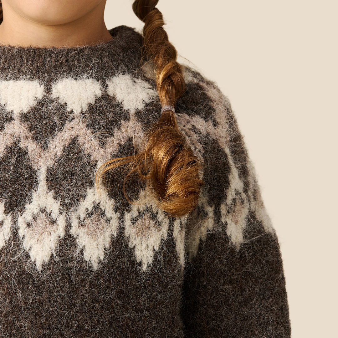 A young girl modeling a chunky knit crewneck sweater with a brown and cream argyle pattern, paired with a black faux-leather mini skirt, against a solid beige background.