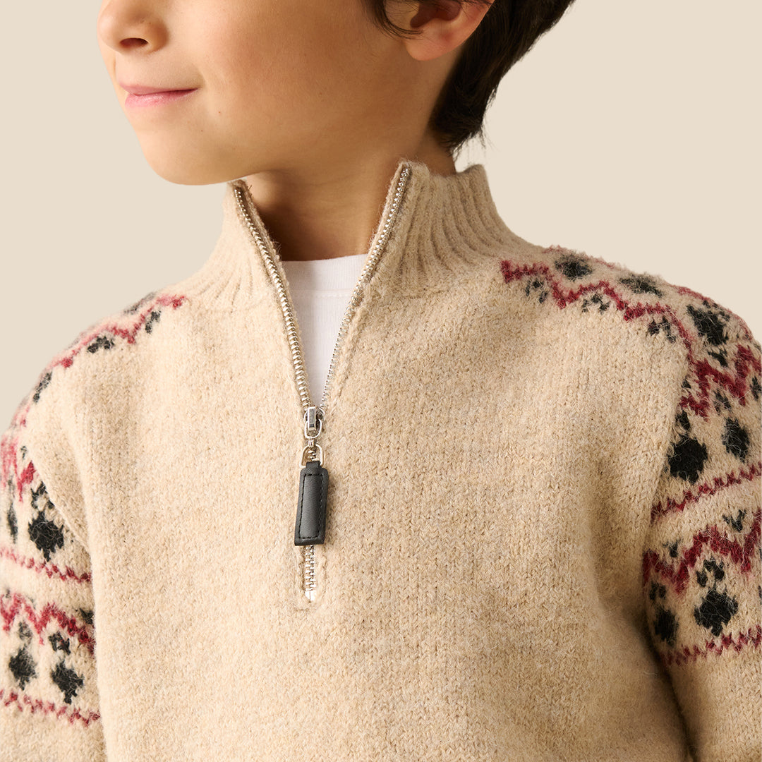 A young boy wearing a beige knit half-zip sweater with red and black Fair Isle patterns on the shoulders and sleeves, set against a solid light beige background.