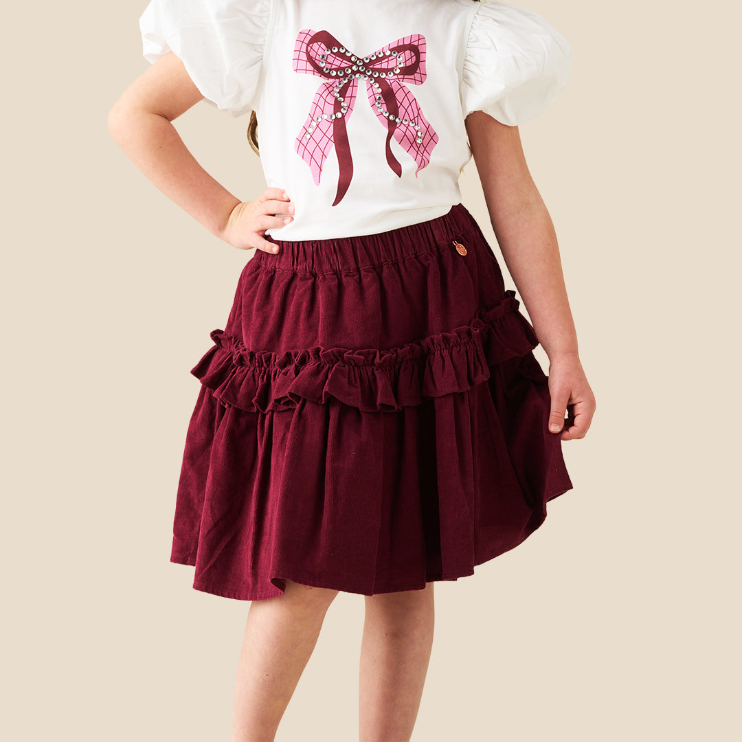 A child wearing a tiered burgundy skirt with ruffles and a white puff-sleeve top featuring a pink plaid bow graphic, photographed against a plain beige background.