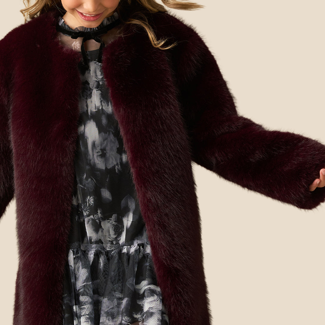 A young girl with blonde hair wearing a plush, dark burgundy faux fur coat over a dark patterned dress, standing against a solid beige background.
