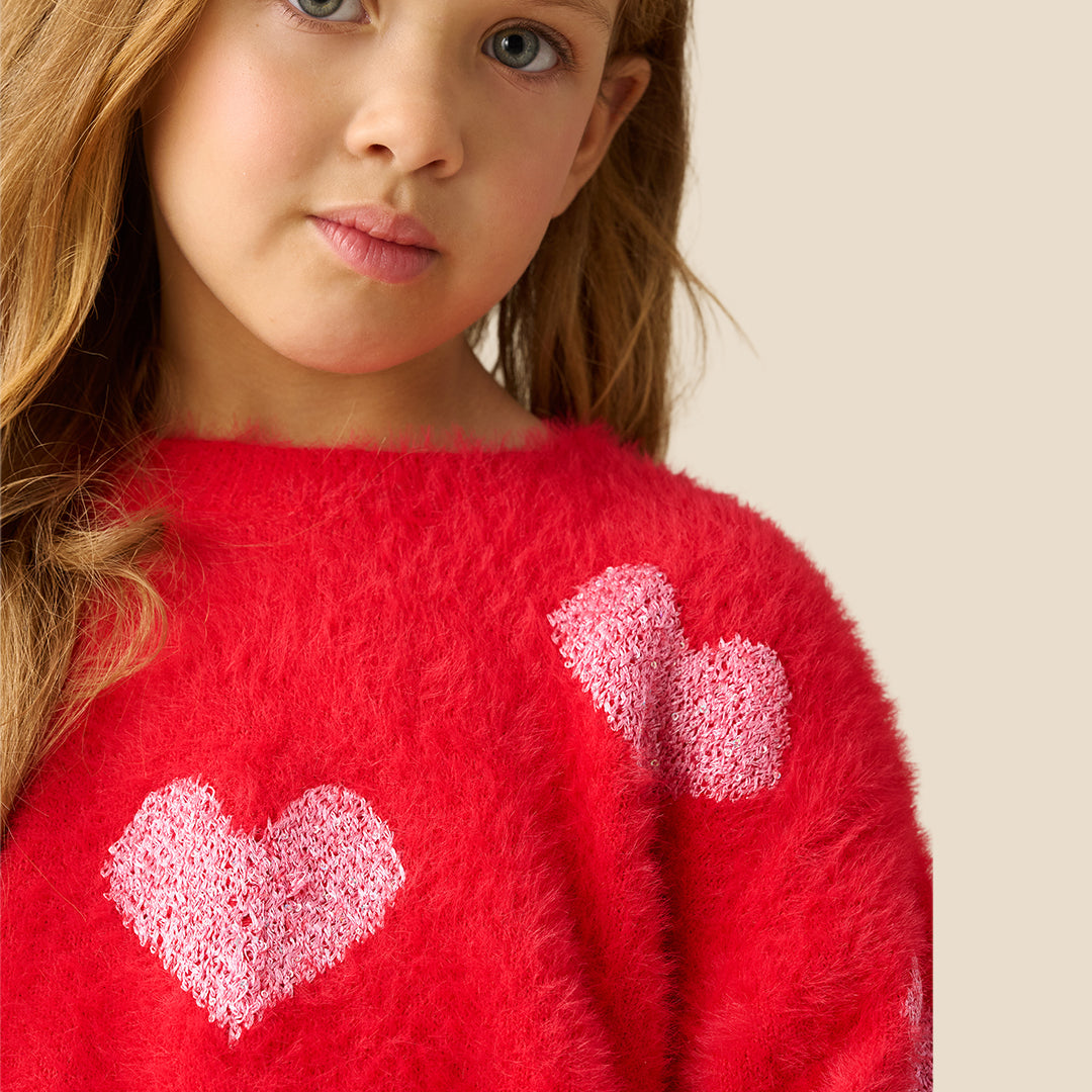 A young girl with long light brown hair wearing a fuzzy red knit sweater with light pink hearts, posing against a solid beige background.