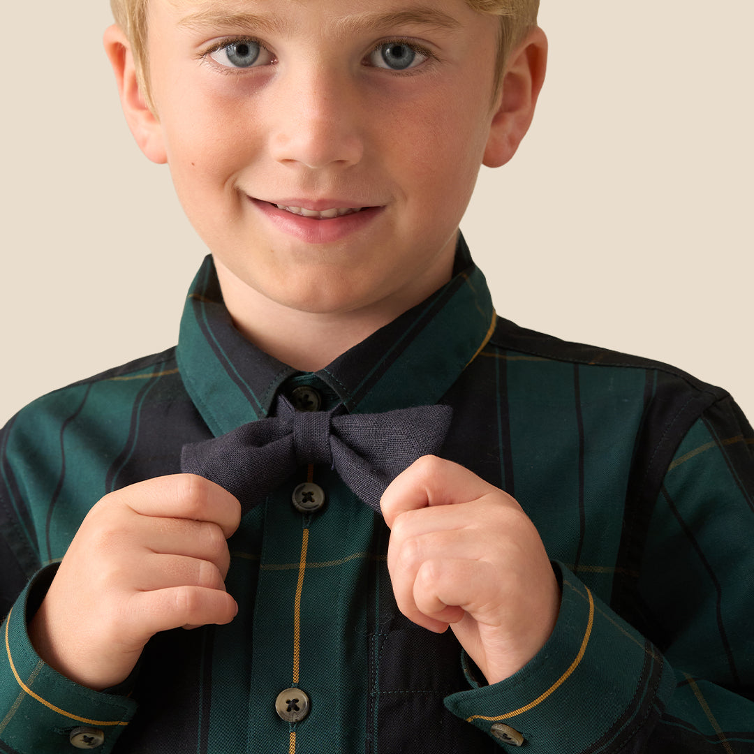A young blond boy wearing a long-sleeve button-up shirt with a dark green and black plaid pattern, a matching dark bow tie, and dark pants, standing against a solid beige background.