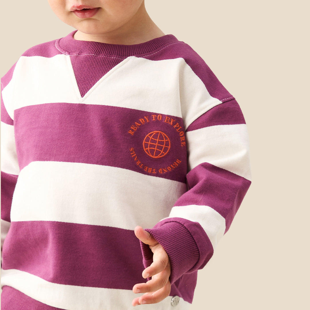 A toddler standing against a solid beige background, wearing a purple and white horizontally striped long-sleeve sweatshirt, dark navy pants, and white sneakers.
