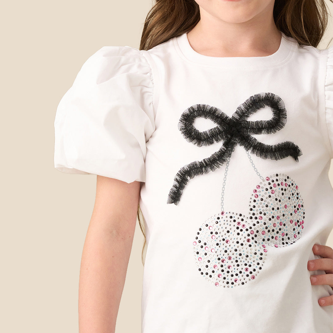 A young girl modeling a white short-sleeve top with puff sleeves and a sequined cherry graphic, paired with black sequined shorts, against a solid beige background.