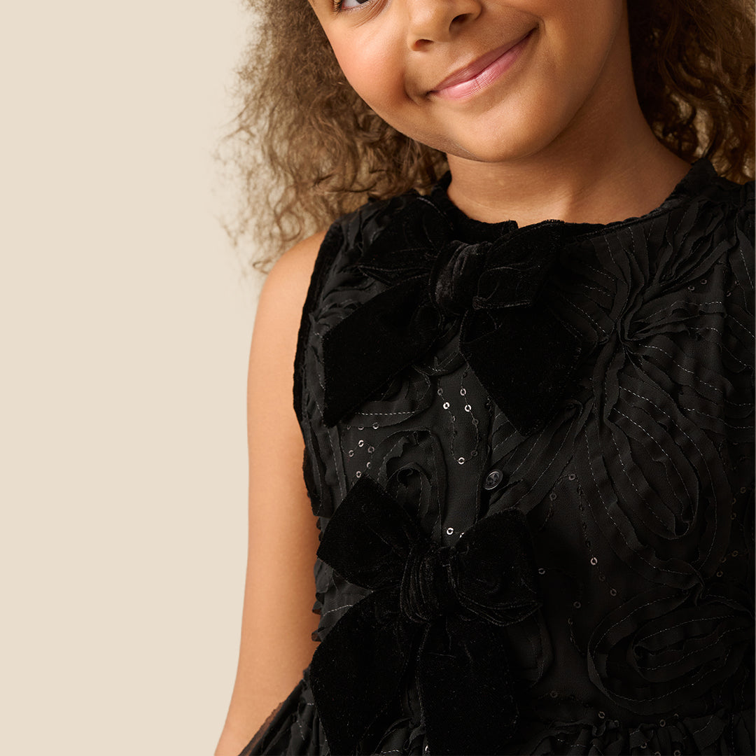 A young girl with curly brown hair wearing a sleeveless black textured dress with a ruffled peplum and bow detail, posing against a solid beige background.