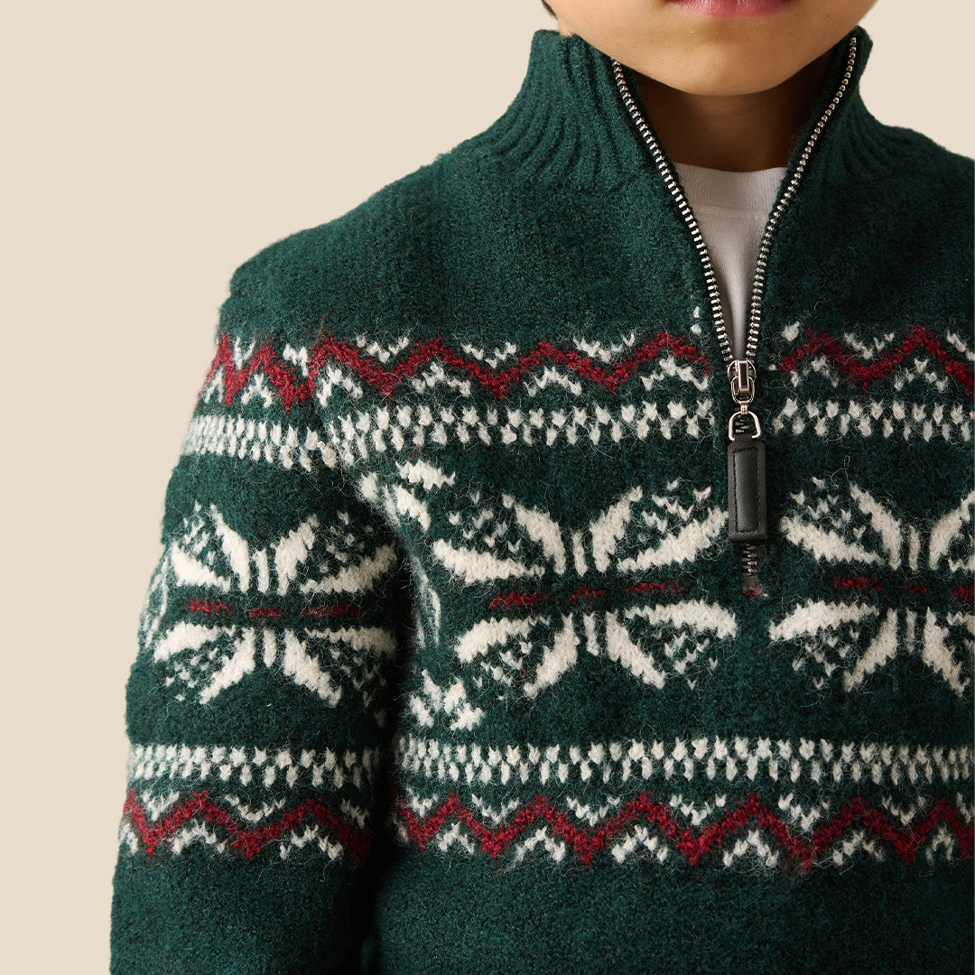 A young boy wearing a dark green knit half-zip sweater with a white and burgundy Fair Isle pattern across the chest and sleeves, paired with dark brown pants, set against a solid beige background.