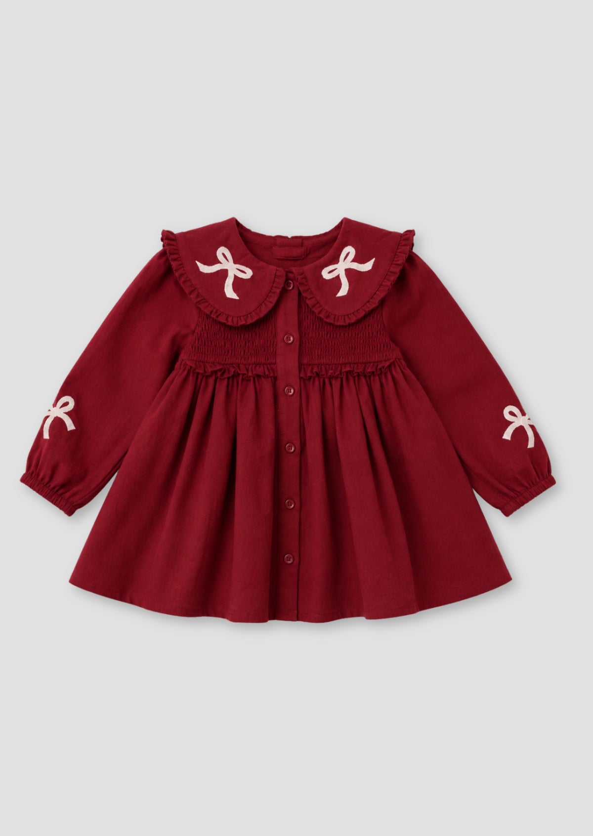 A toddler girl wearing a burgundy corduroy dress with a large white ruffled collar decorated with bows, white tights, and beige ballet flats, standing against a solid beige background.