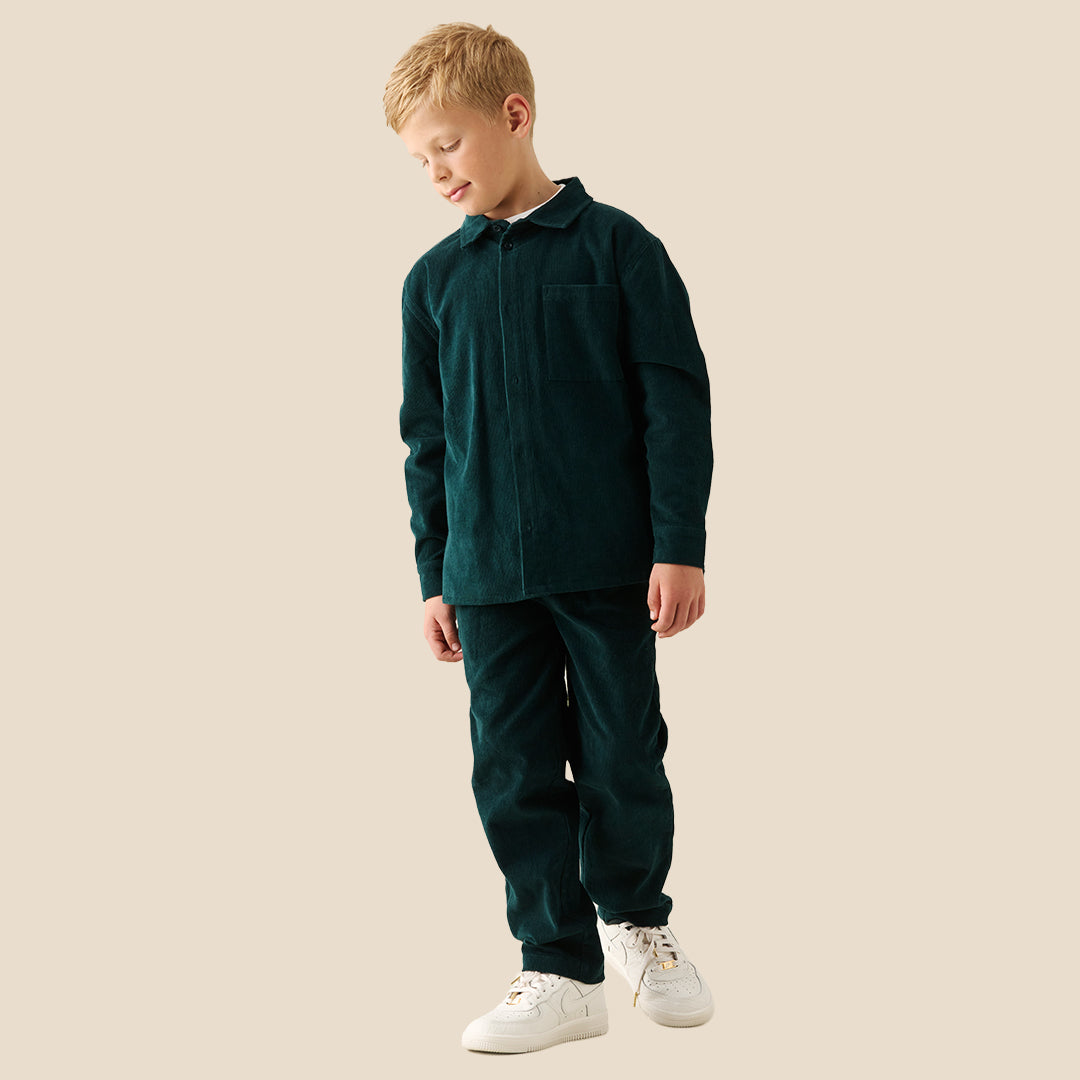A young boy with blonde hair wearing a dark forest green corduroy button-up overshirt over a white t-shirt and matching green pants, standing against a solid beige background.