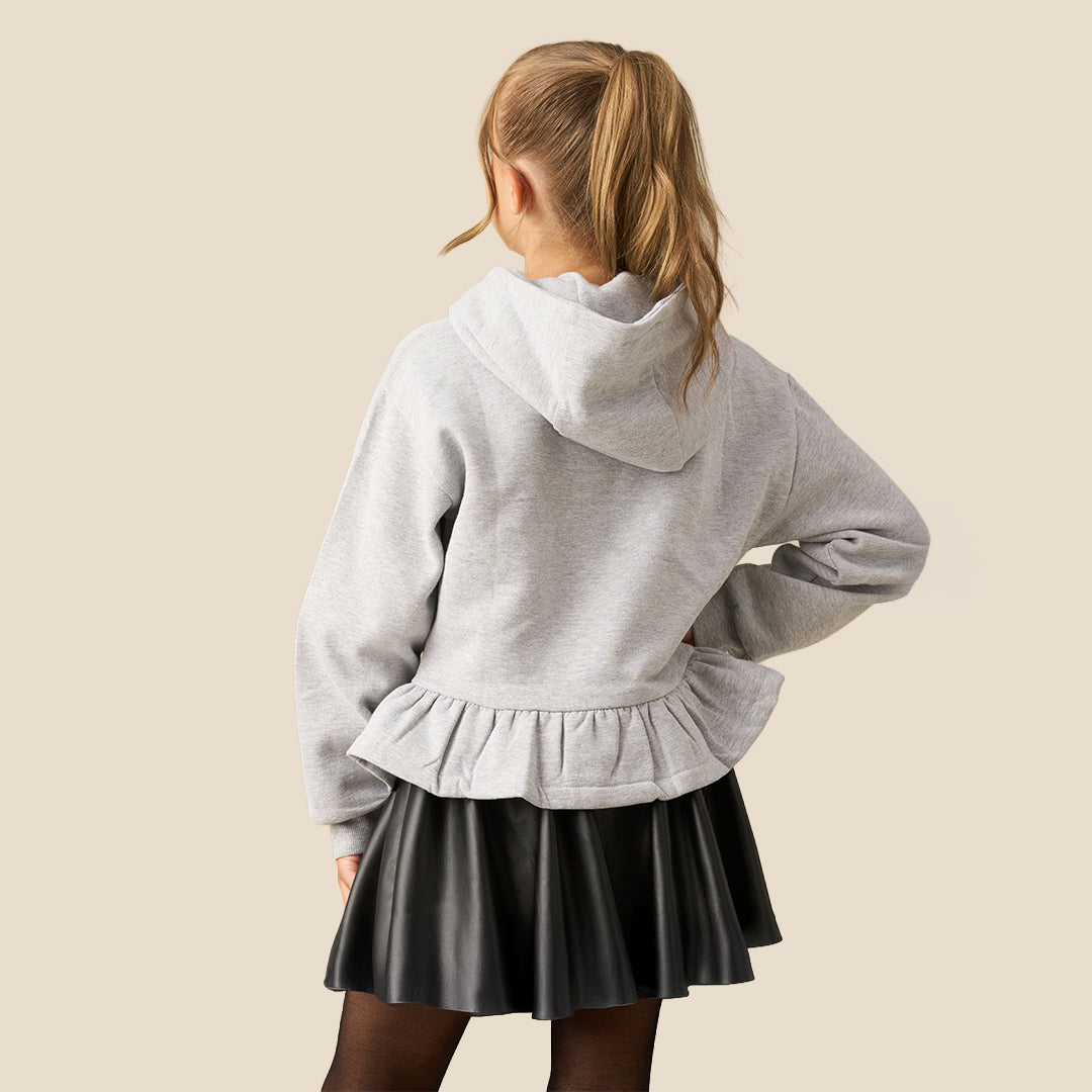 A young girl with her hair in a ponytail models a light gray heathered hoodie featuring a large black bow graphic on the front and a ruffled hem, paired with a black faux-leather skirt, against a solid beige background.