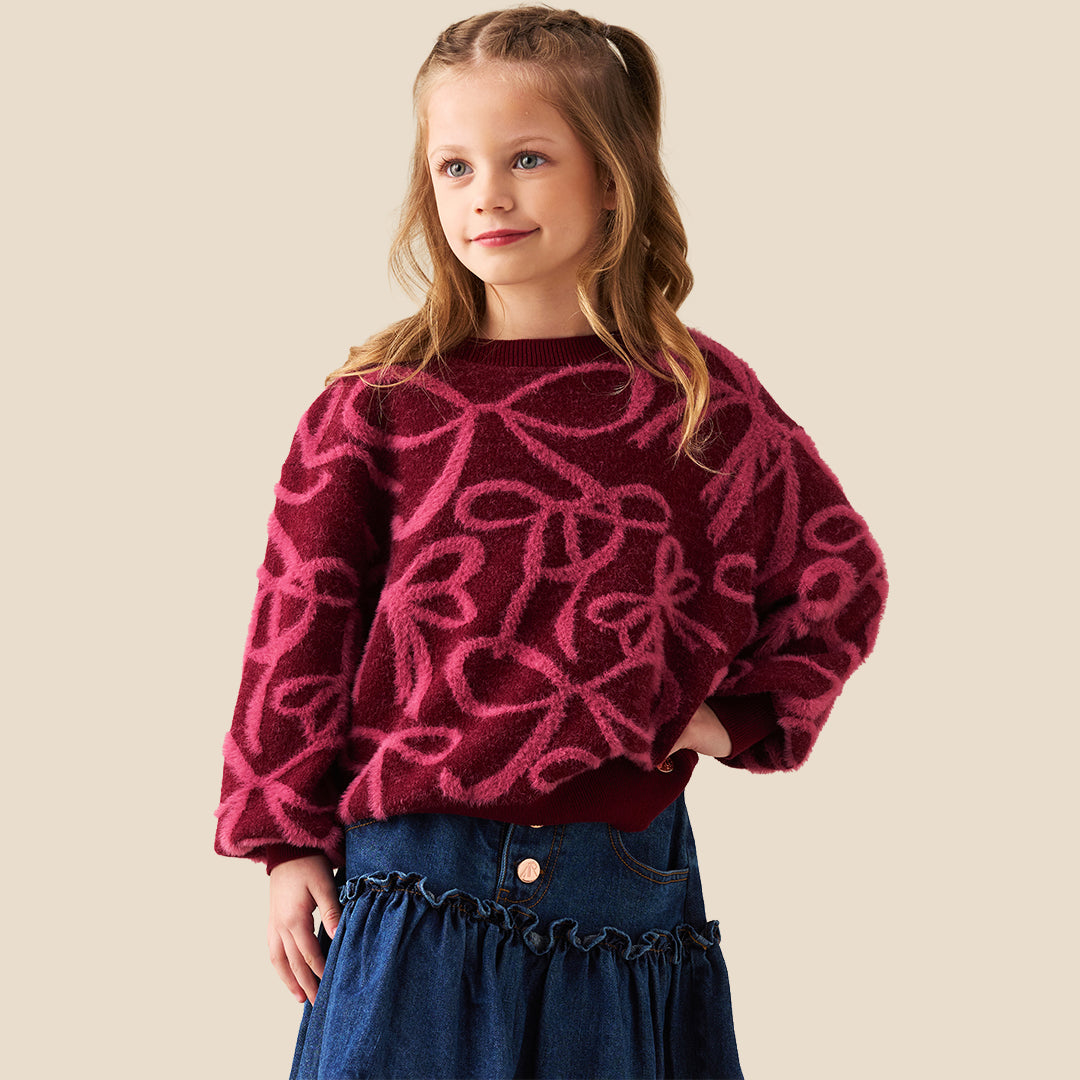 A young girl with blonde hair in a ponytail wearing a fuzzy dark red long-sleeve sweater with a pink bow pattern, paired with a blue denim skirt, against a solid beige background.