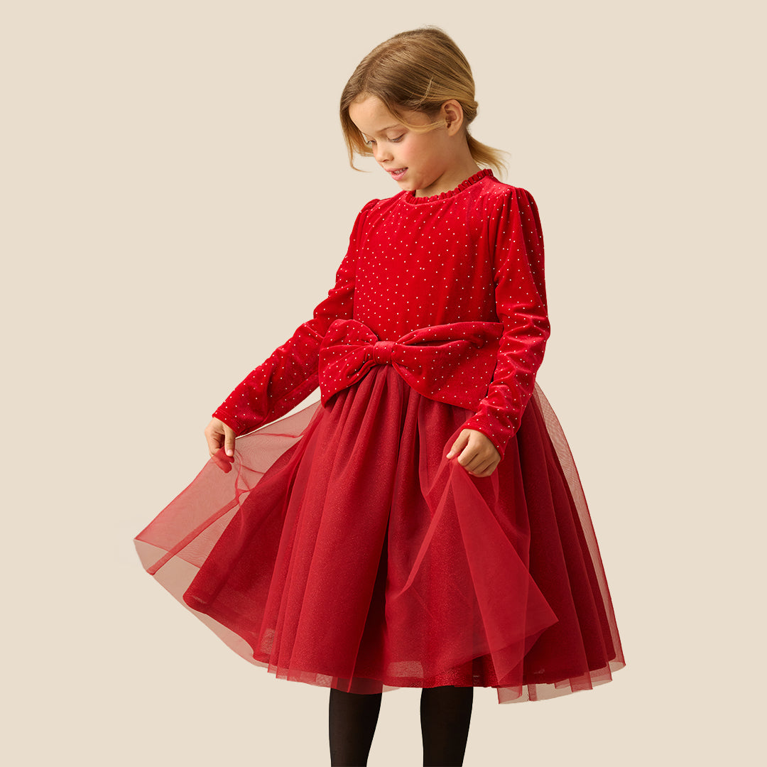 A young girl wearing a festive red dress with long sleeves, tiny gold polka dots, a matching red bow at the waist, and a tulle skirt, set against a solid beige background.