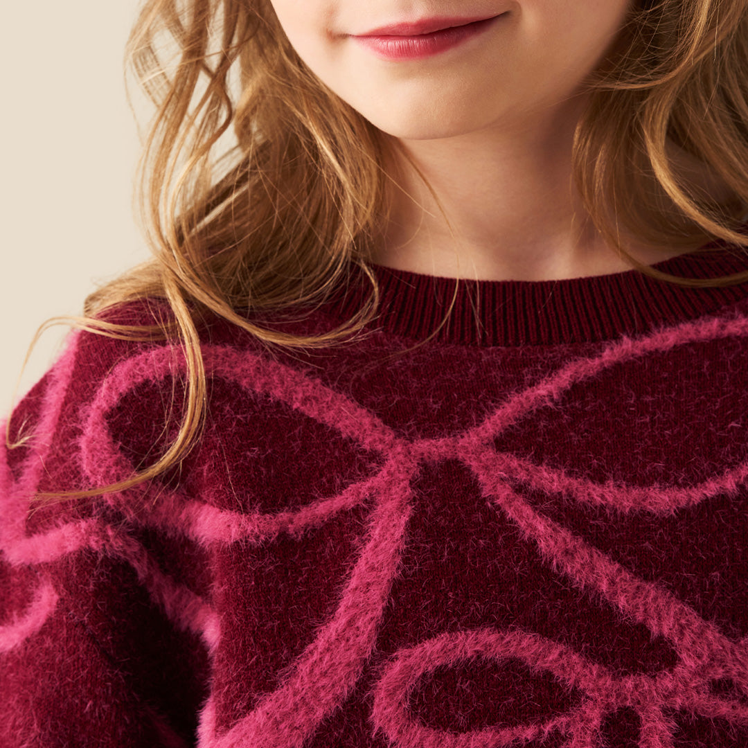 A young girl with blonde hair in a ponytail wearing a fuzzy dark red long-sleeve sweater with a pink bow pattern, paired with a blue denim skirt, against a solid beige background.