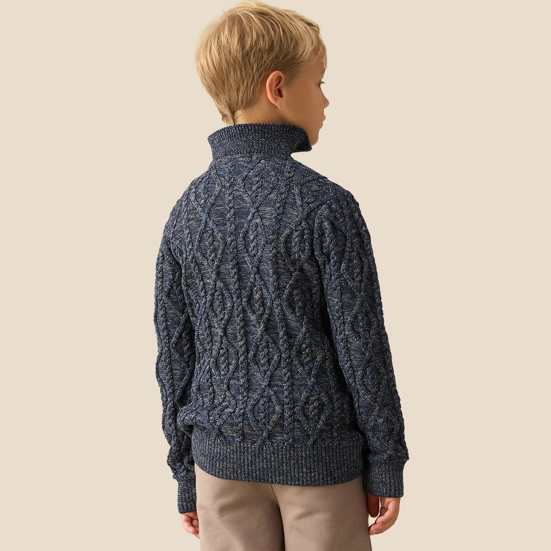 A young boy with blonde hair wearing a navy blue cable-knit mock-neck sweater and beige trousers, standing against a solid beige background.