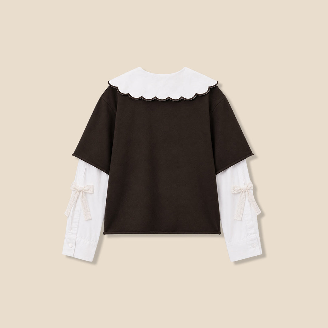 A young girl with wavy light brown hair is wearing a dark brown short-sleeve top with a large white ruffled collar featuring black cursive embroidery, layered over a white long-sleeve shirt, paired with brown and black leopard-print pants, against a solid beige background.