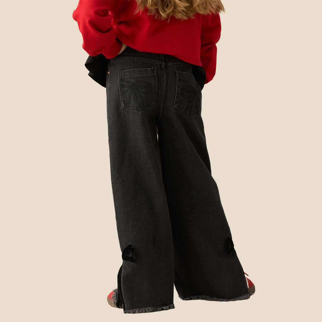 A person wearing black wide-leg denim jeans with side tie details and raw frayed hems, styled with a red bow-tie sweatshirt and red sneakers, against a solid beige background.