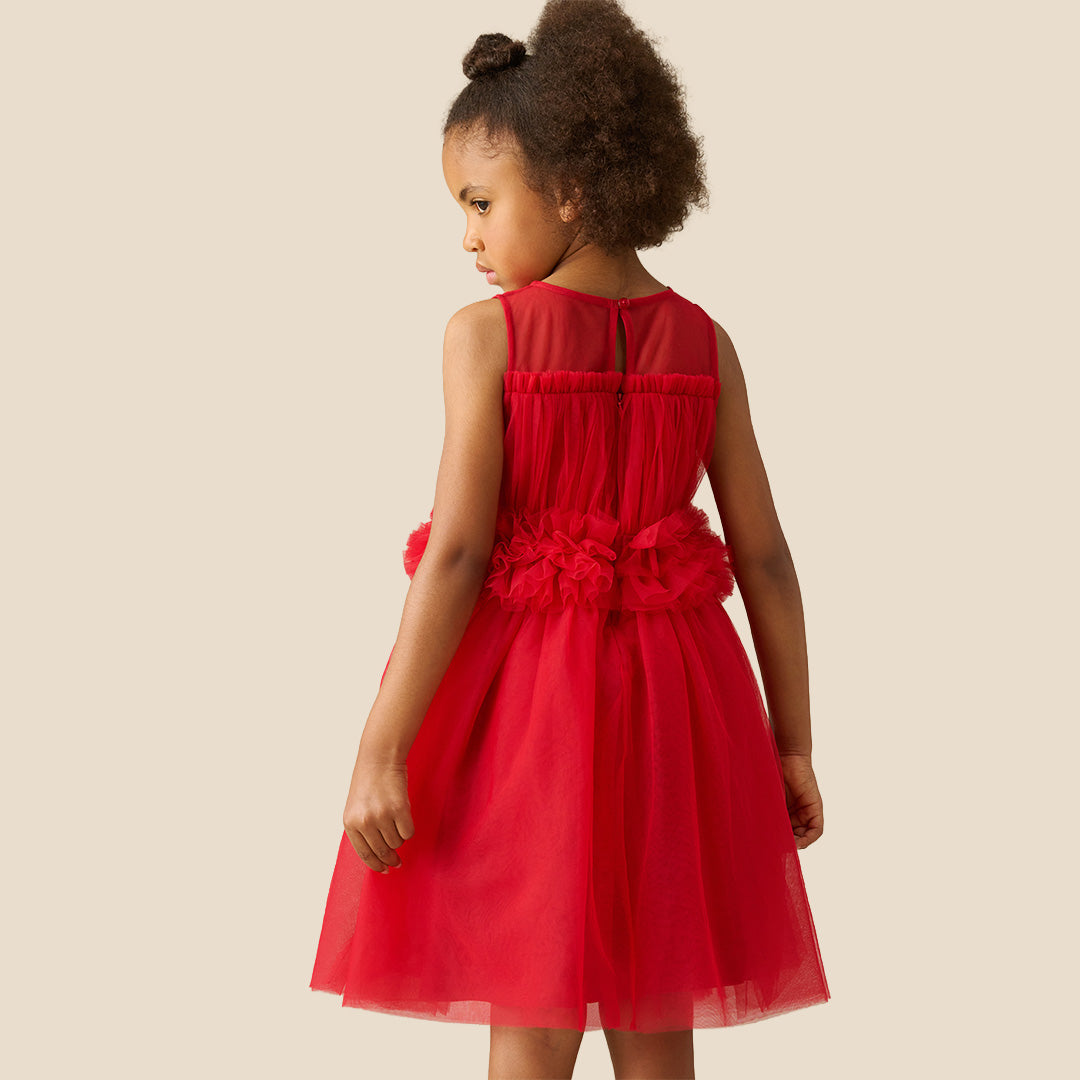 A young girl with curly brown hair wearing a sleeveless bright red tulle dress with a ruffled waistband, standing against a solid beige background.