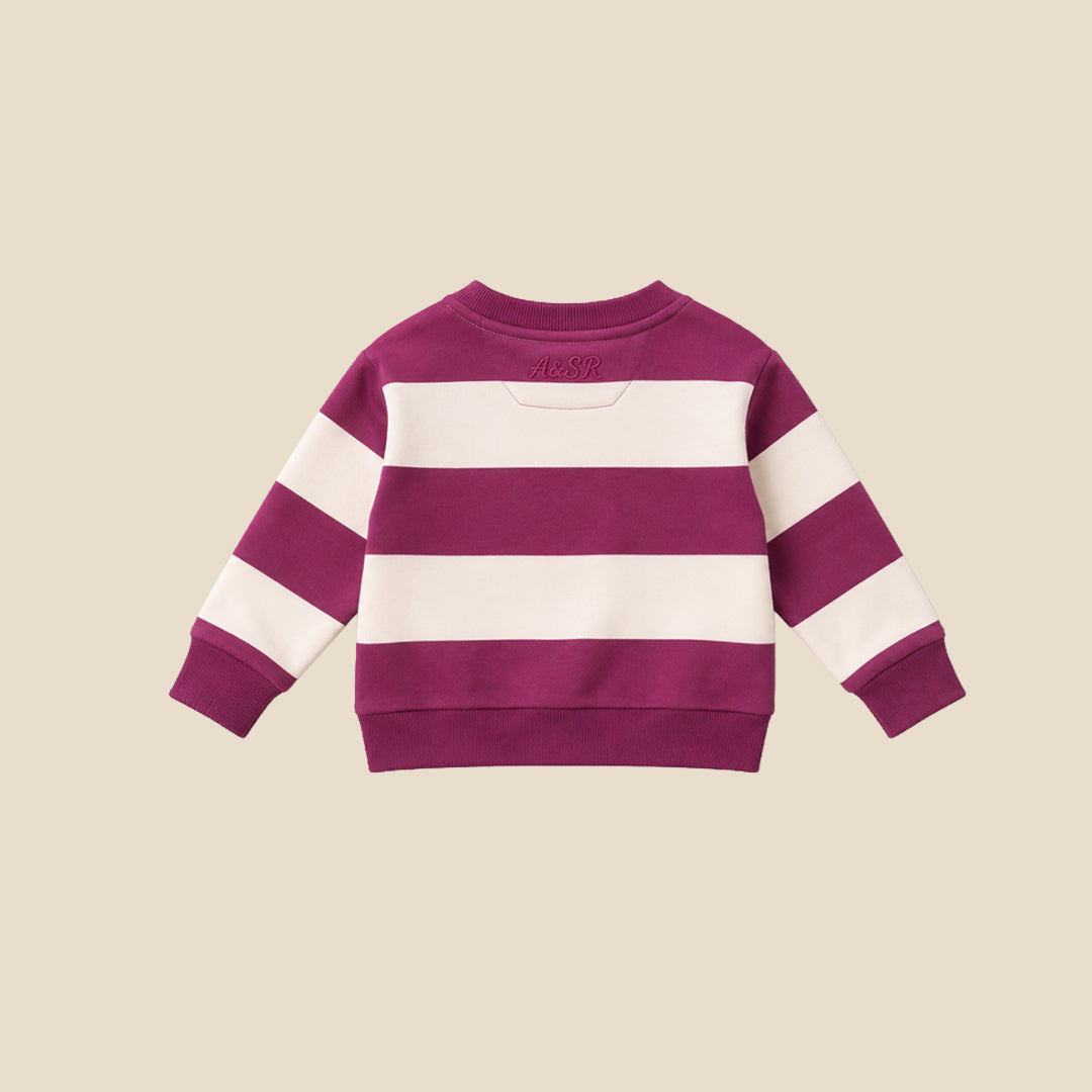 A toddler standing against a solid beige background, wearing a purple and white horizontally striped long-sleeve sweatshirt, dark navy pants, and white sneakers.
