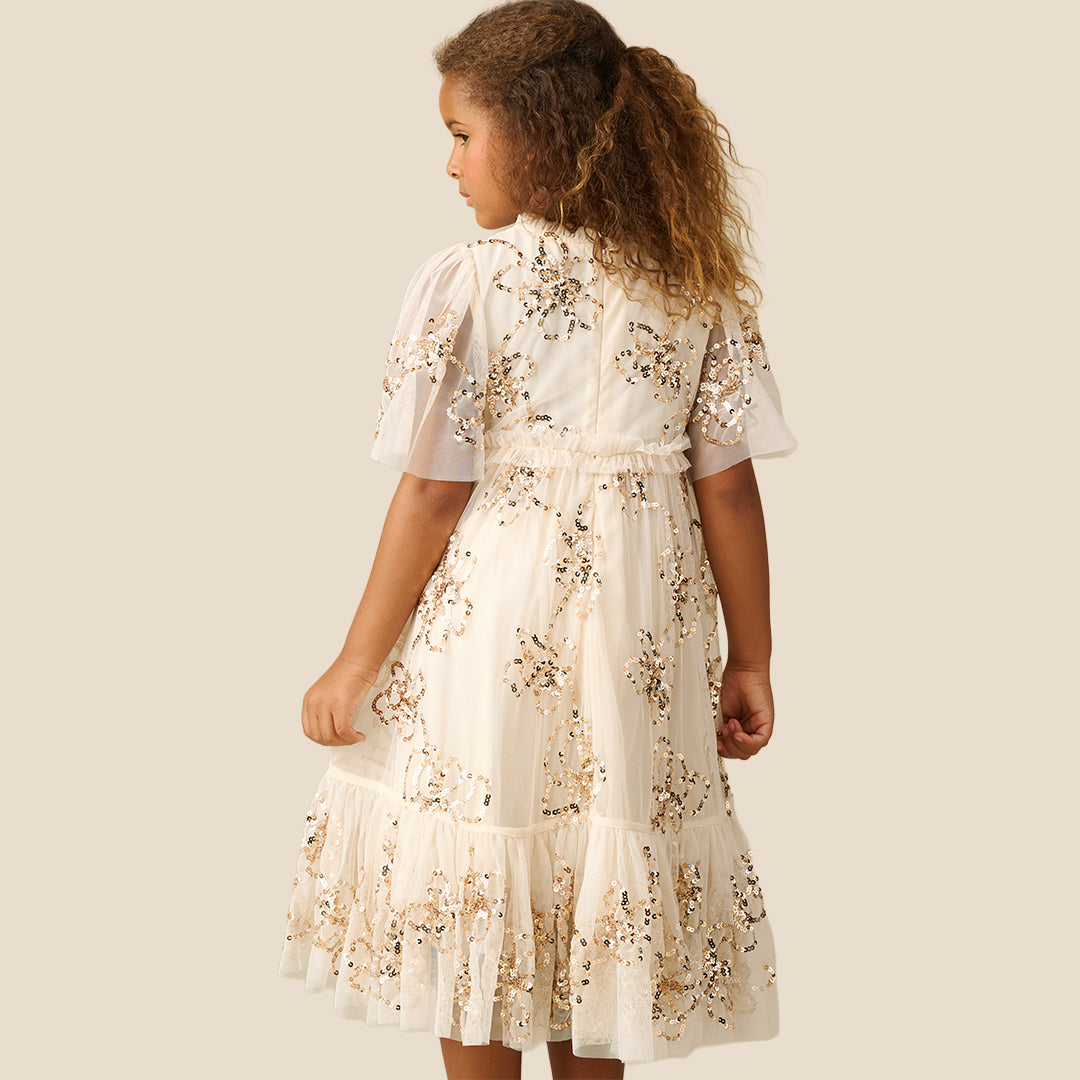 A young girl with curly brown hair wearing a tiered cream dress decorated with gold sequins and floral embroidery, standing against a solid beige background.