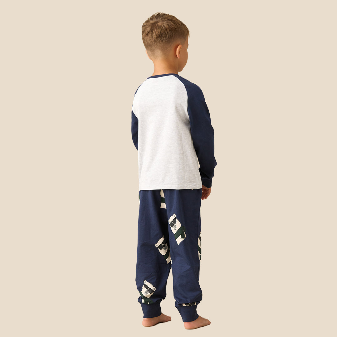 A young boy modeling a two-piece pajama set against a solid beige background. The set features a white long-sleeve top with navy blue raglan sleeves and a cartoon bear graphic wearing sunglasses and a green scarf, paired with navy blue pants decorated with patchwork-style graphic details.