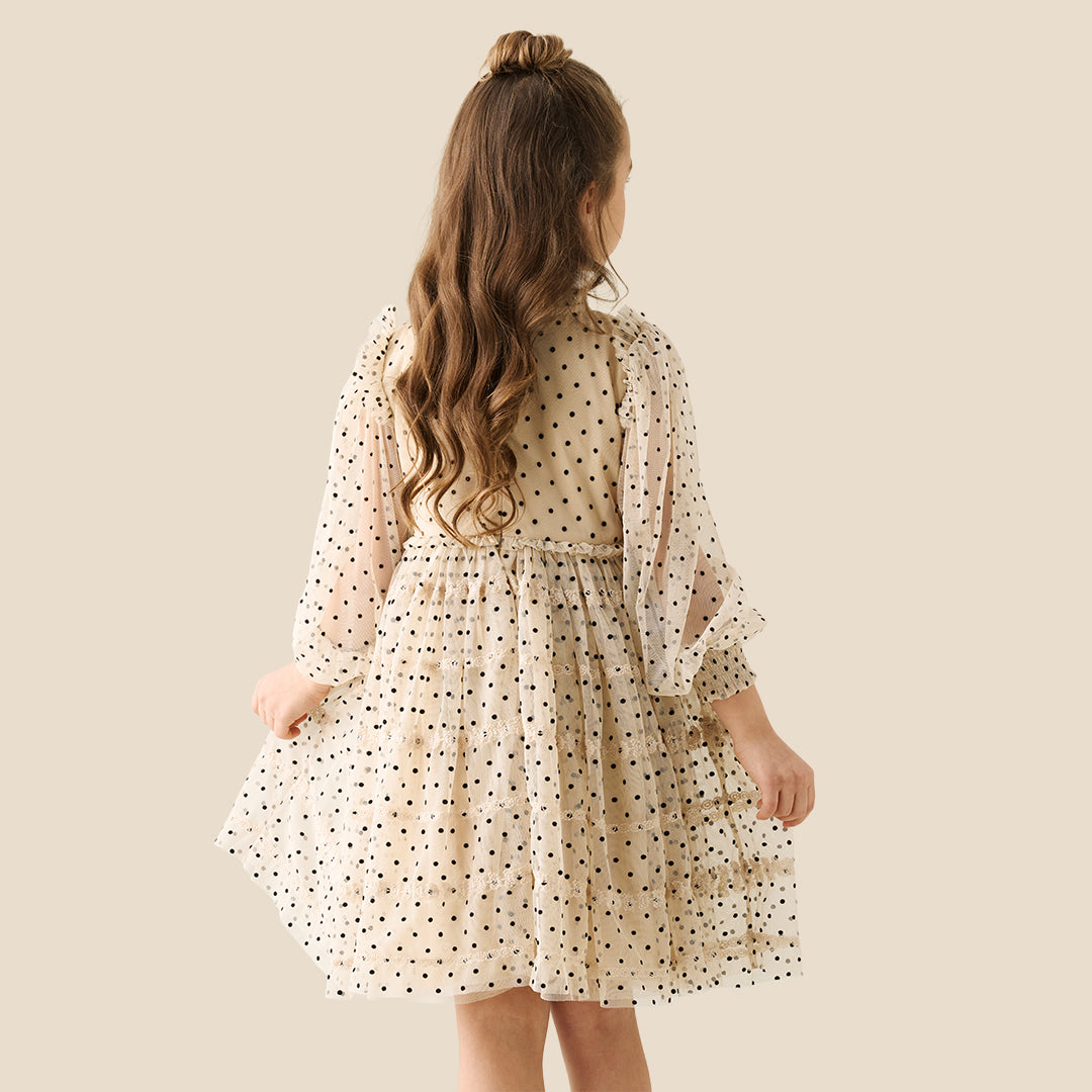 A young girl with her hair in a bun wears a cream-colored, long-sleeved tiered dress with black polka dots, standing against a solid beige background.