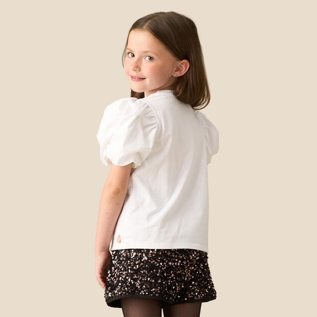 A young girl modeling a white short-sleeve top with puff sleeves and a sequined cherry graphic, paired with black sequined shorts, against a solid beige background.