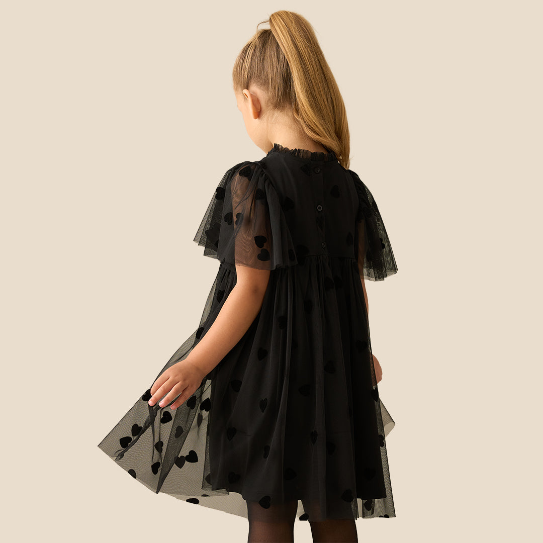A young girl with her hair in a ponytail models a black tulle dress with heart patterns and sheer flutter sleeves, set against a solid beige background.