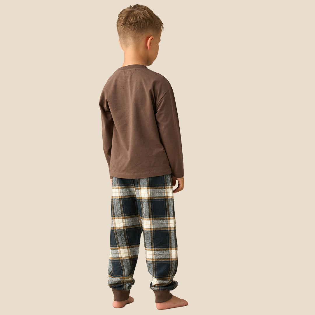 A young boy modeling a two-piece pajama set consisting of a brown long-sleeve shirt with a plaid chest pocket and matching plaid pants, set against a solid beige background.
