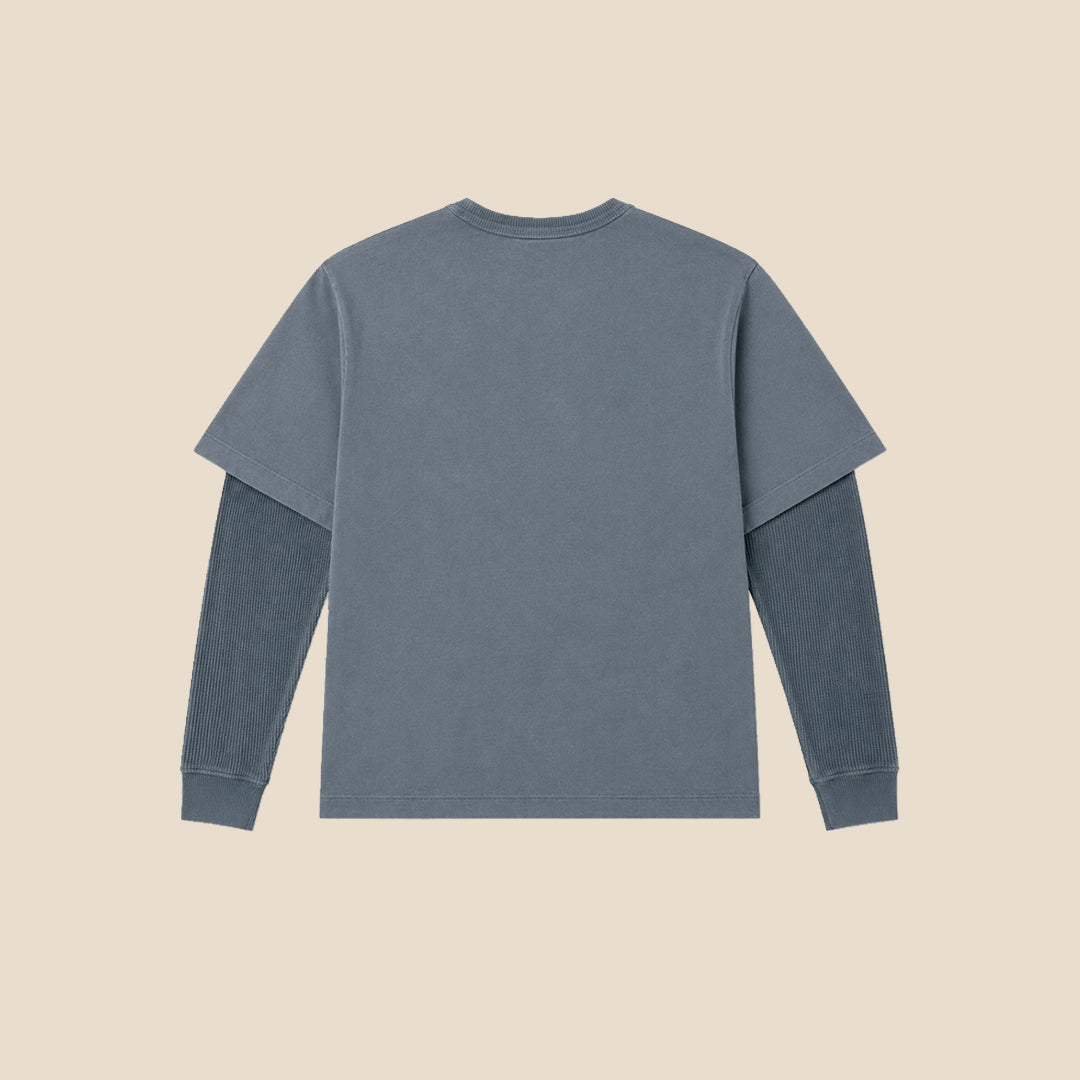 A young boy modeling a washed slate blue long-sleeve crewneck shirt with a chest pocket, paired with beige cargo pants, against a solid beige background.