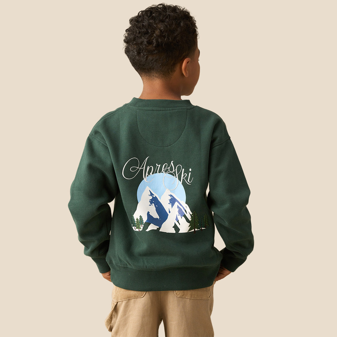 A young boy with curly dark hair wearing a dark green crewneck sweatshirt with a small white graphic on the left chest and khaki pants, posing against a solid light beige background.
