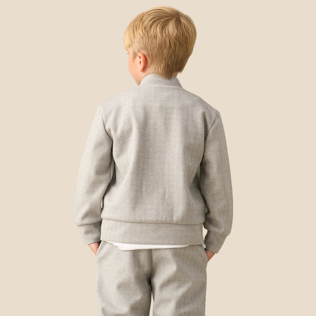A young boy with blond hair wearing a light gray zip-up fleece jacket and matching gray sweatpants, standing against a solid beige background.