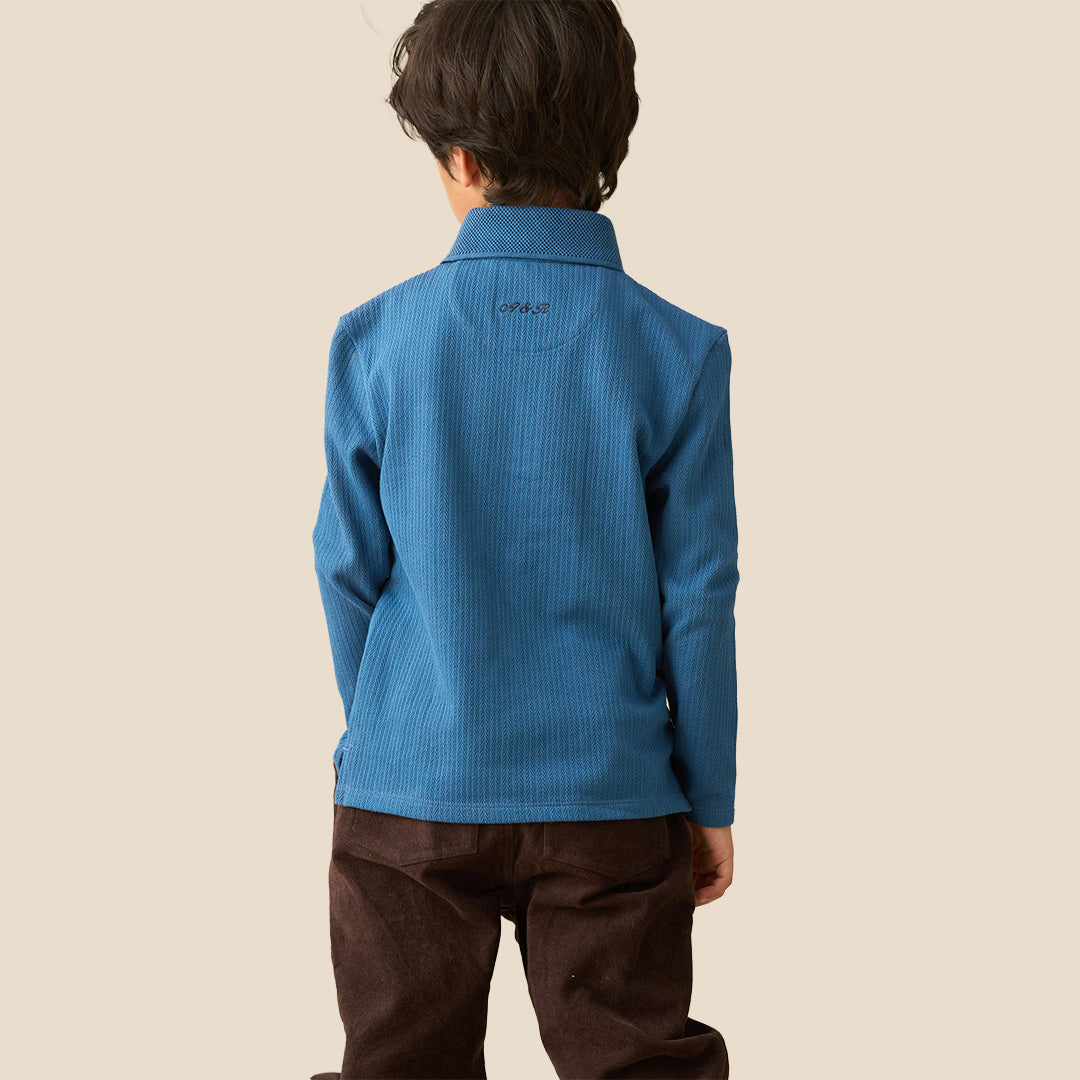 A young boy wearing a blue ribbed long-sleeve polo shirt and brown pants, standing against a solid beige background.