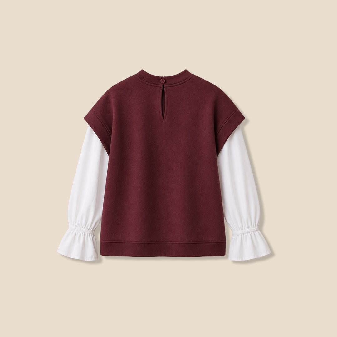 A young girl with blonde pigtails wearing a maroon cropped t-shirt with white sleeves and 'C'EST LA VIE paris' printed in white, layered over a white long-sleeve blouse with ruffled cuffs, and blue jeans, against a solid beige background.