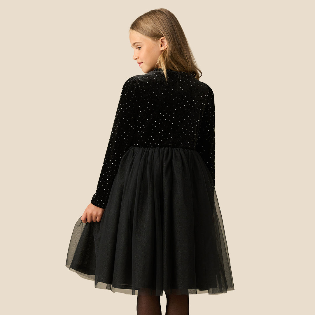 A young girl with blonde hair wearing a black long-sleeve dress with a polka-dot velvet bodice, a sheer black tulle skirt, and a matching bow belt, posing against a solid beige background.