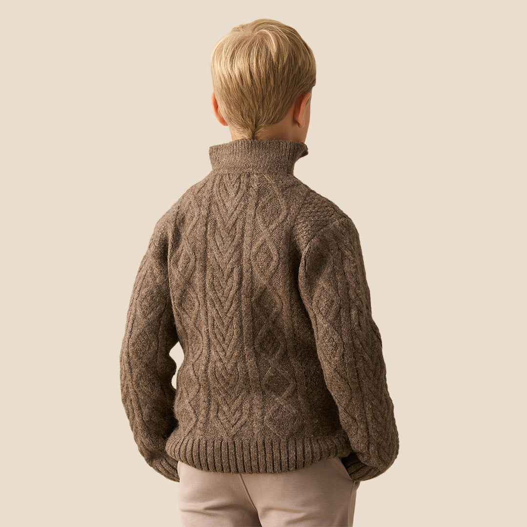 A child wearing a brown cable-knit mock-neck sweater and light pink trousers, standing indoors near a white wall and window. Another child in a brown-and-white plaid shirt is partially visible in the foreground.