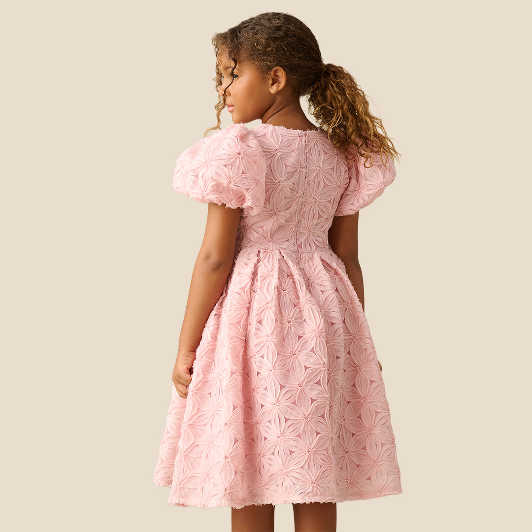 A young girl with curly brown hair wearing a light pink textured dress with puffed short sleeves and a full skirt, standing against a solid beige background.