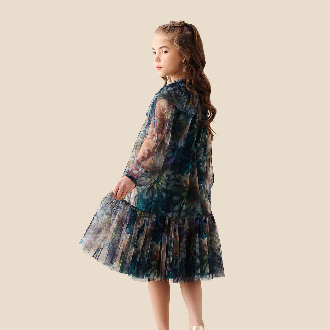A young girl modeling a sheer, tiered tulle dress with a large ruffled collar and a blue, green, and purple floral watercolor pattern, set against a solid beige background.