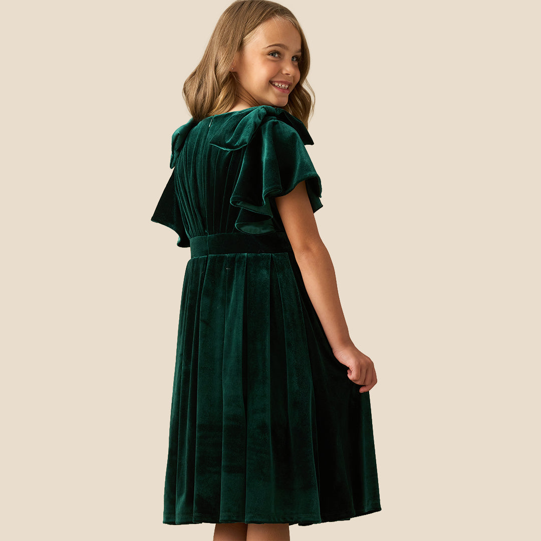 A young girl wearing a dark green velvet dress with ruffled short sleeves, a matching waist belt, and a pleated skirt, standing against a solid beige background.