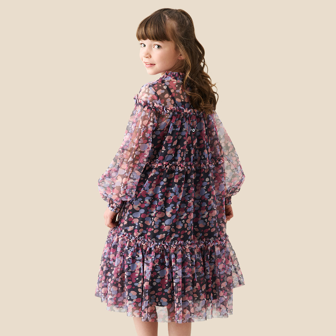A young girl with curly hair stands on a checkered porch wearing a long-sleeved tiered dress with a pink, purple, and blue floral pattern. She is posing behind a black wrought-iron fence.