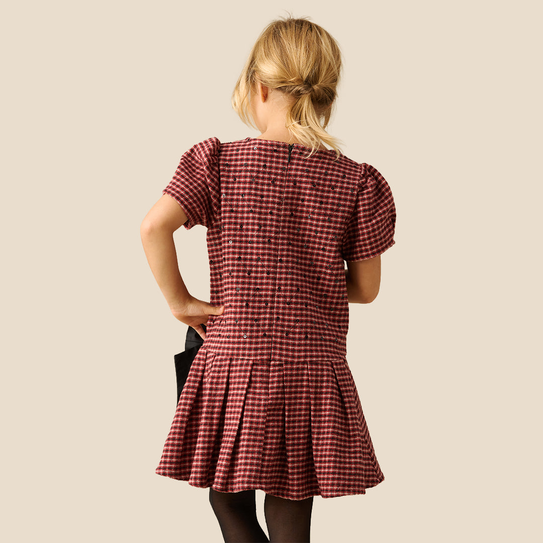 A young girl smiling and posing in a red and white gingham dress with a large black bow at the waist, set against a solid beige background.