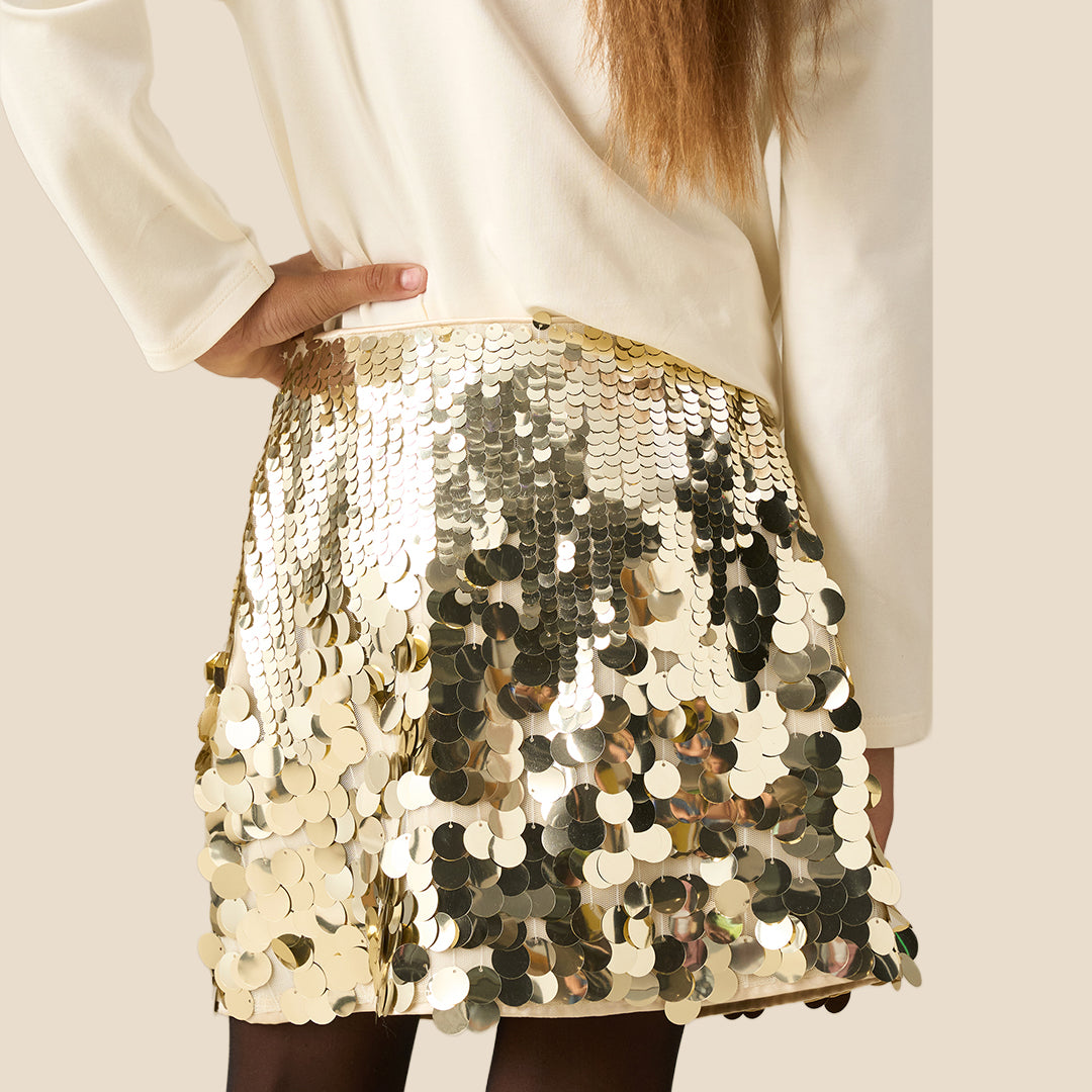 A close-up of a person wearing a shiny gold sequin mini skirt with a matching gold chain belt, paired with a cream-colored top and black tights, against a solid beige background.