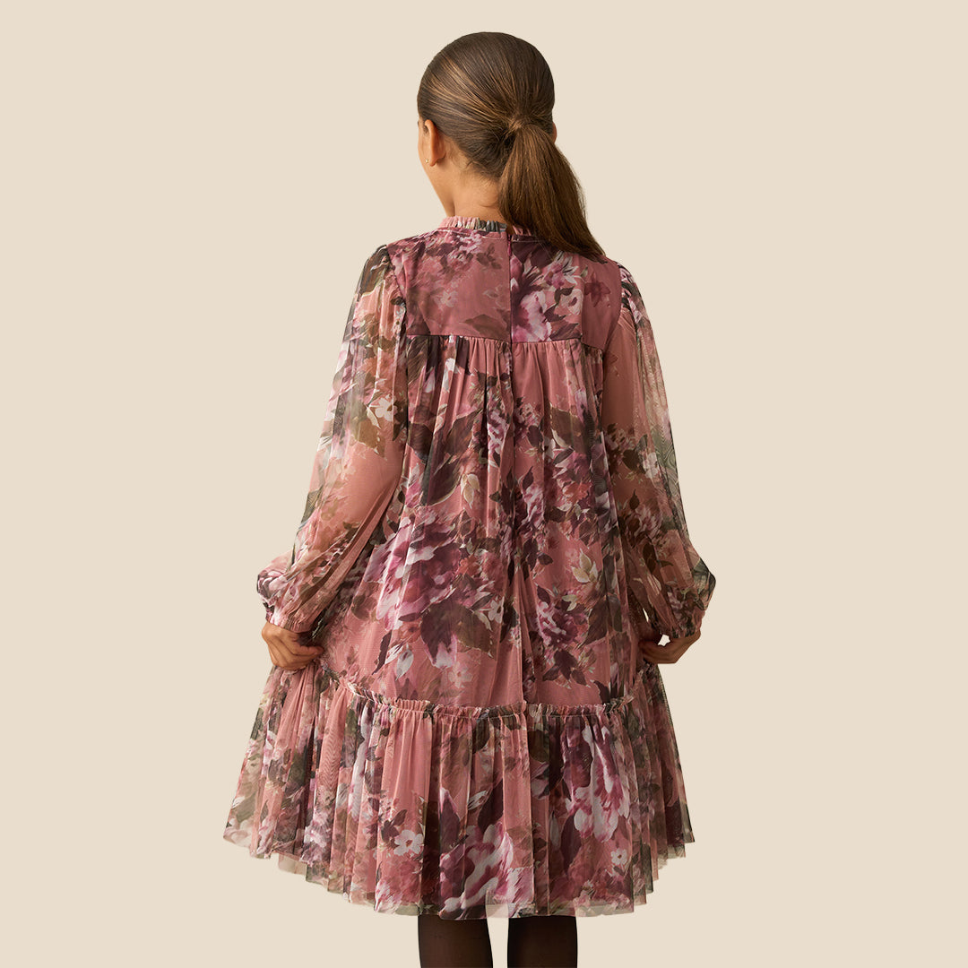 A young girl with her hair in a ponytail models a long-sleeve tiered dress with a dusty rose and brown floral print against a solid beige background.