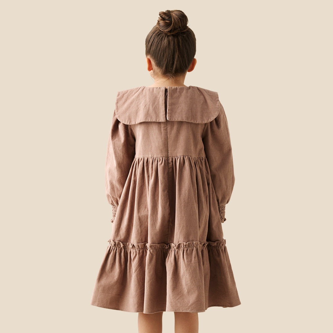 A young girl with her hair in a bun wears a brown corduroy dress with a large ruffled collar and bow, standing against a solid beige background.