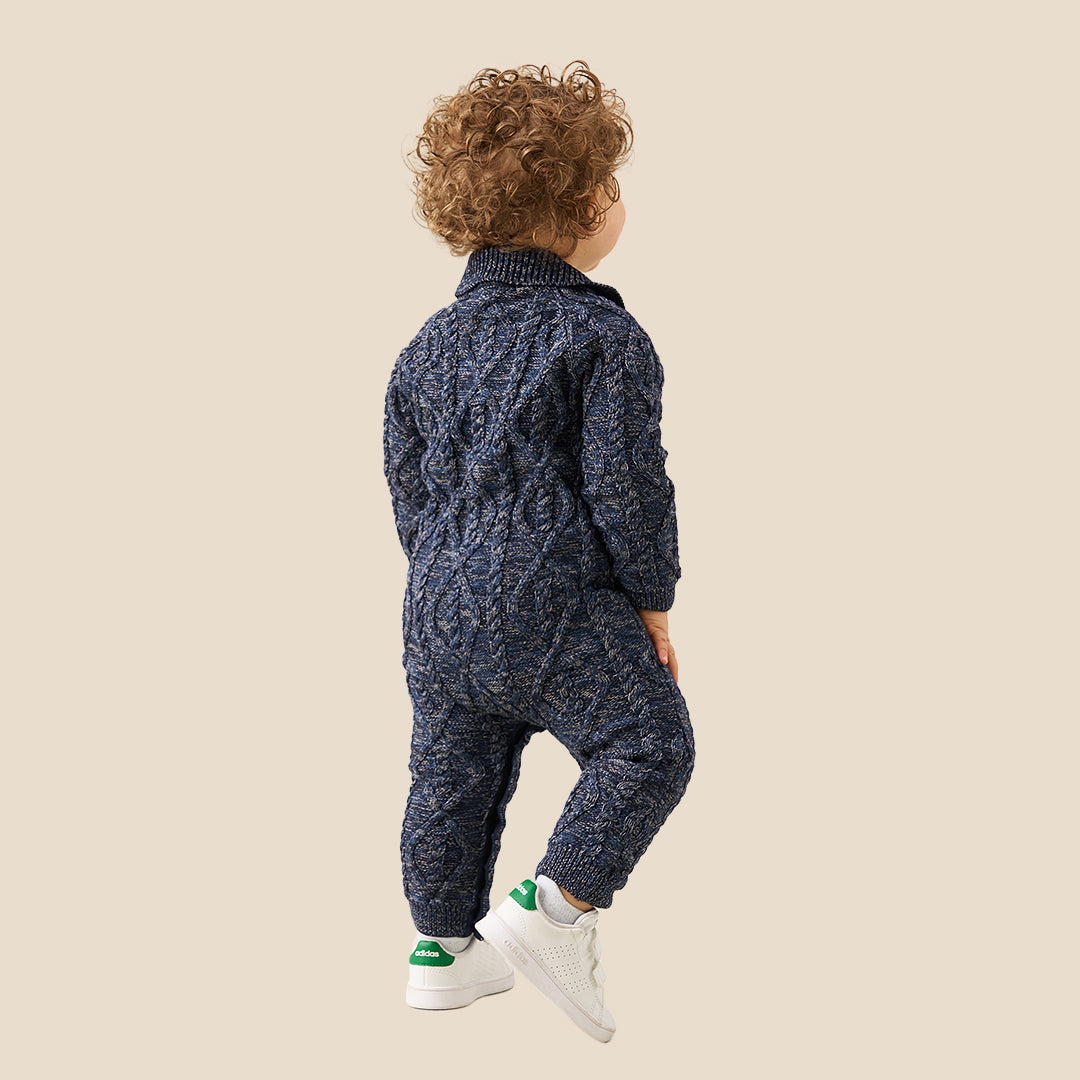 A baby with curly light brown hair wearing a navy blue cable-knit one-piece jumpsuit and white shoes, standing against a solid beige background.