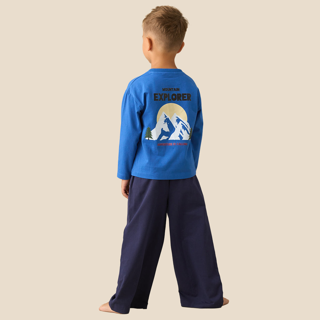 A young boy modeling a two-piece pajama set consisting of a bright blue long-sleeve shirt and navy blue wide-leg pants with pink text on the leg, set against a solid beige background.