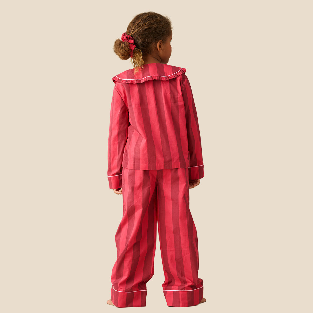 A young girl modeling a two-piece pajama set with vertical pink and red stripes, featuring a ruffled collar and white piping details, set against a solid beige background.