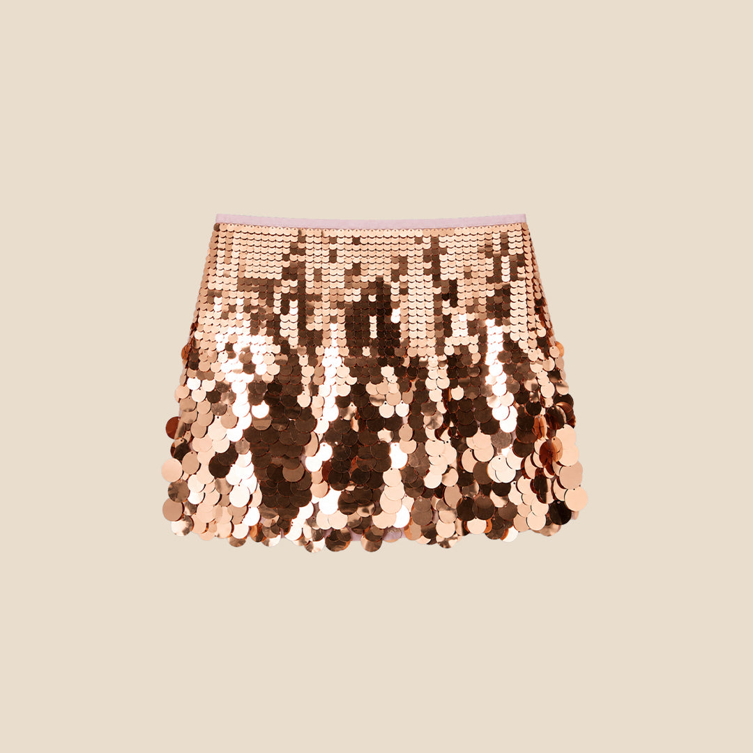 A child wearing a white graphic T-shirt and a short skirt covered in large, reversible sequins that shift between rose gold and bronze, photographed against a plain beige studio background.