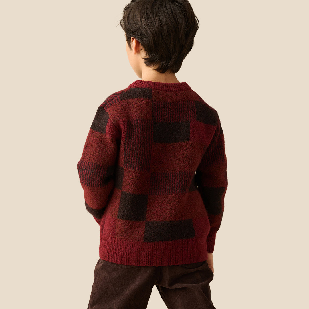 A young boy wearing a textured knit sweater with a dark red and black checkered pattern, paired with dark brown pants, set against a solid light beige background.